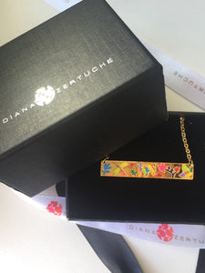 Golden artisanal necklace with colorful floral design displayed in an elegant black box with the logo 'Diana Zertuche' embossed on the lid and matching branded ribbon