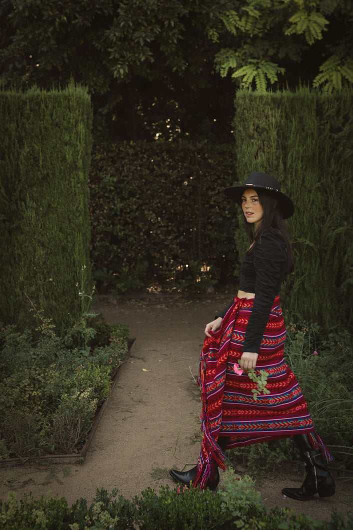 model walking  in a garden wearing a red rebozo as a skirt and a black hat, boots and top